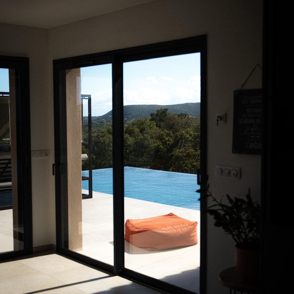 Depuis l’intérieur de la villa, la baie coulissante ouverte sur la piscine à débordement, le maquis et les collines.