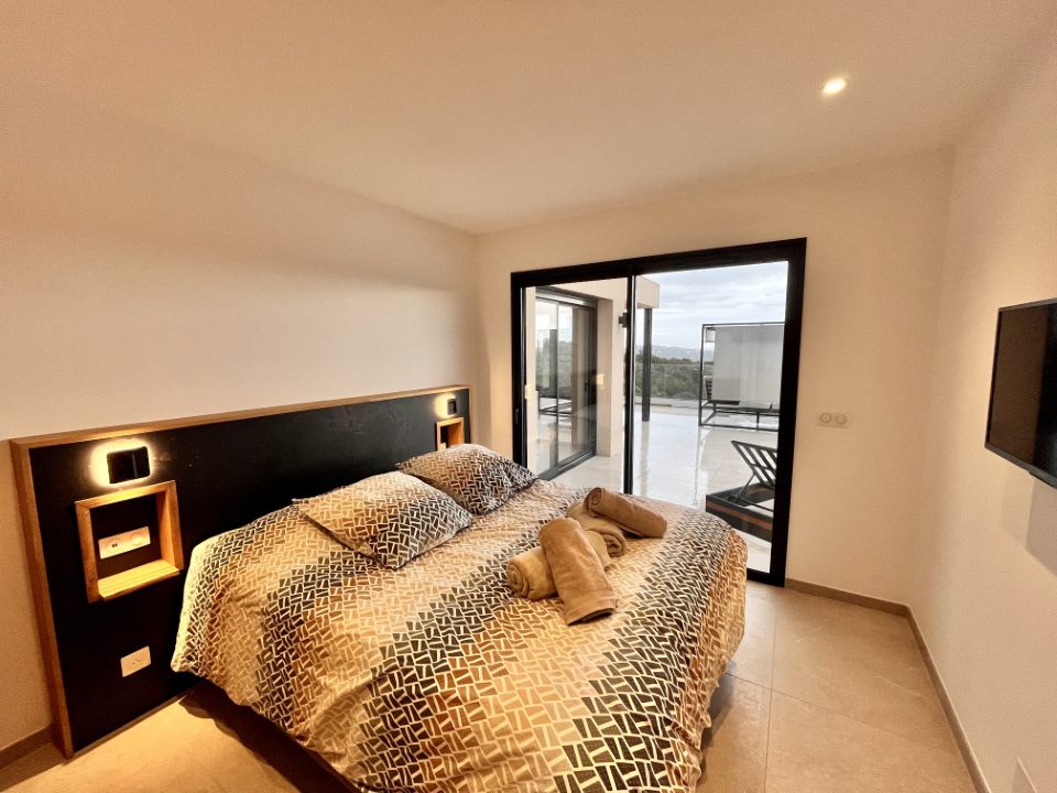 Chambre 1 : lit double, tête de lit avec niches en chêne et liseuses intégrées, baie coulissante vers la terrasse.